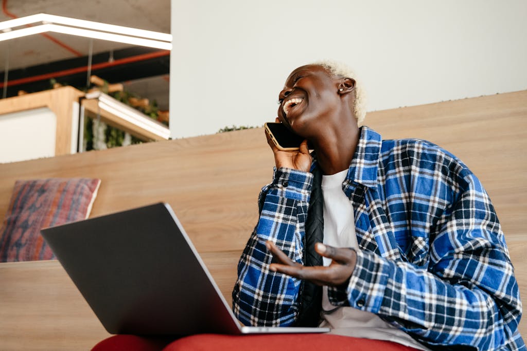 Cheerful black woman with laptop talking on smartphone